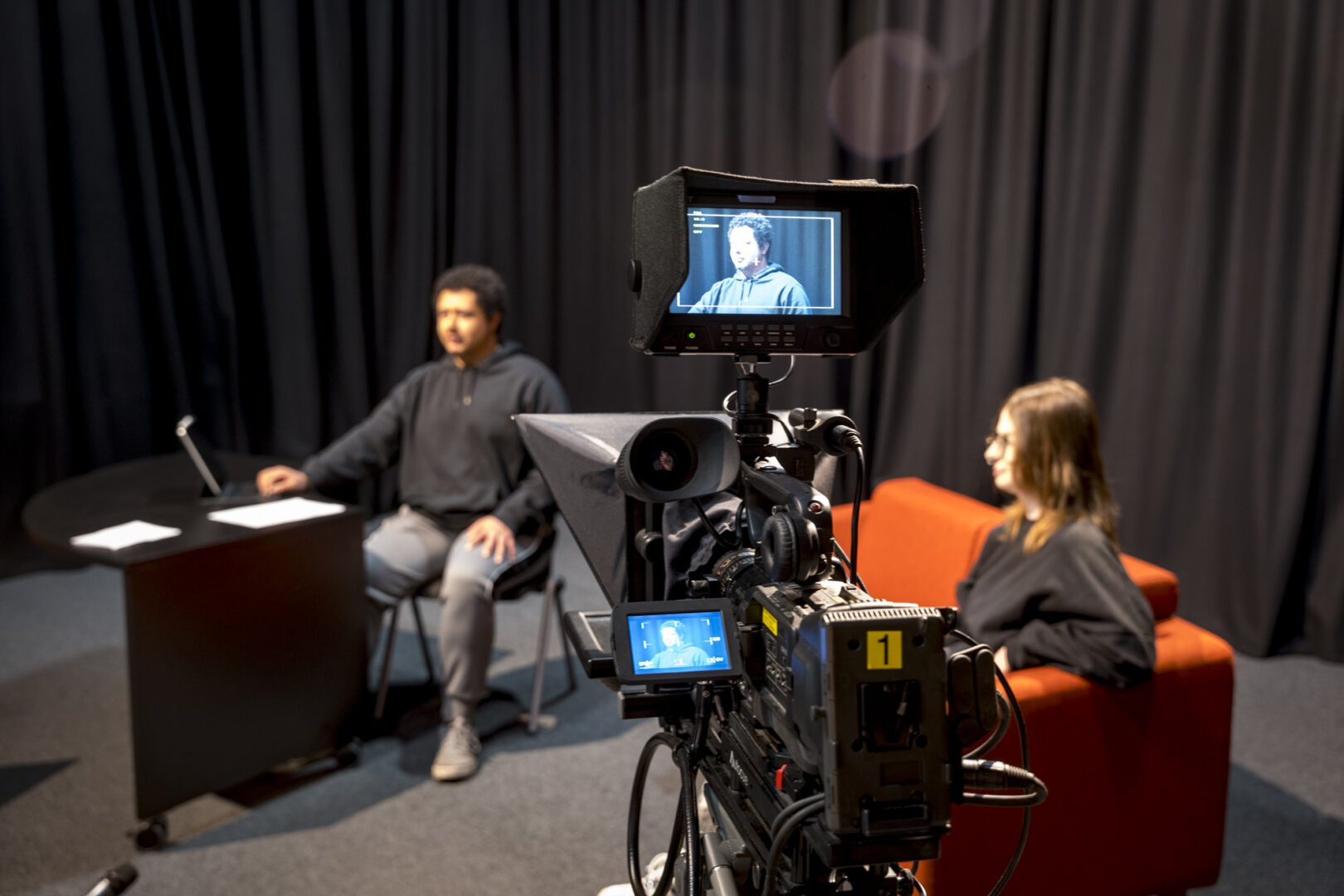 creative media production students sat in the television studio, one student is sat at a desk being filmed while the other sits on a couch
